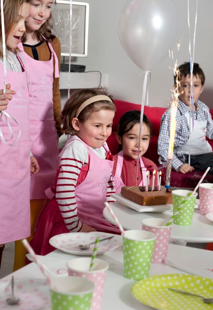 Niños con delantales rosas celebrando con un pastel, velas y fuente luminosa.