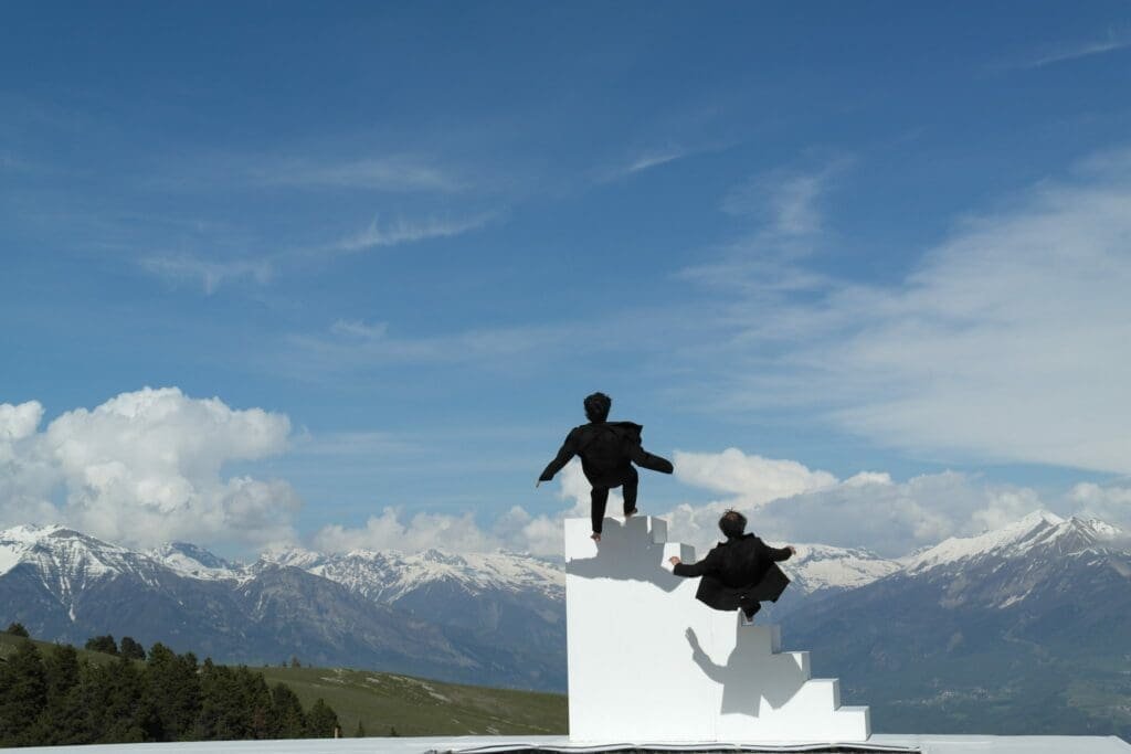 Dos bailarines negros en una escalera blanca ante montañas nevadas, espectáculo al aire libre.