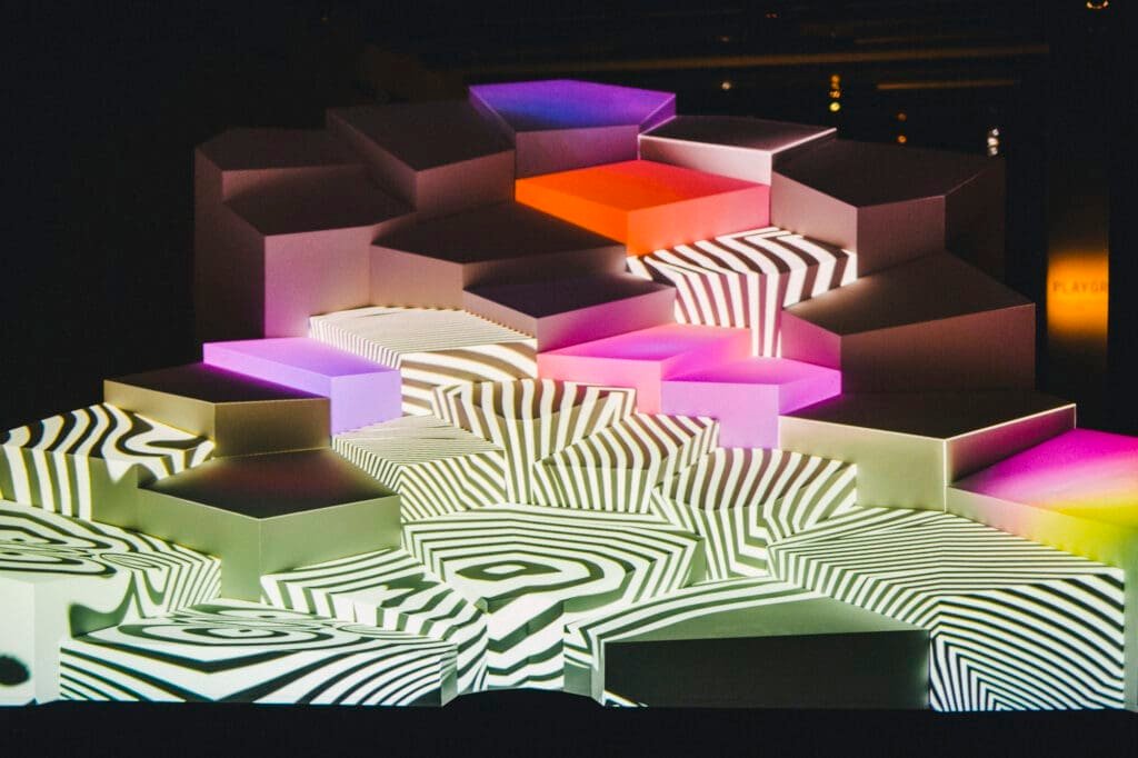 Installation géométrique avec projection de motifs zébrés et blocs lumineux colorés.