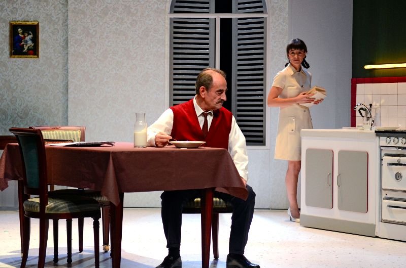 Theater scene: man eating, woman standing in a kitchen.