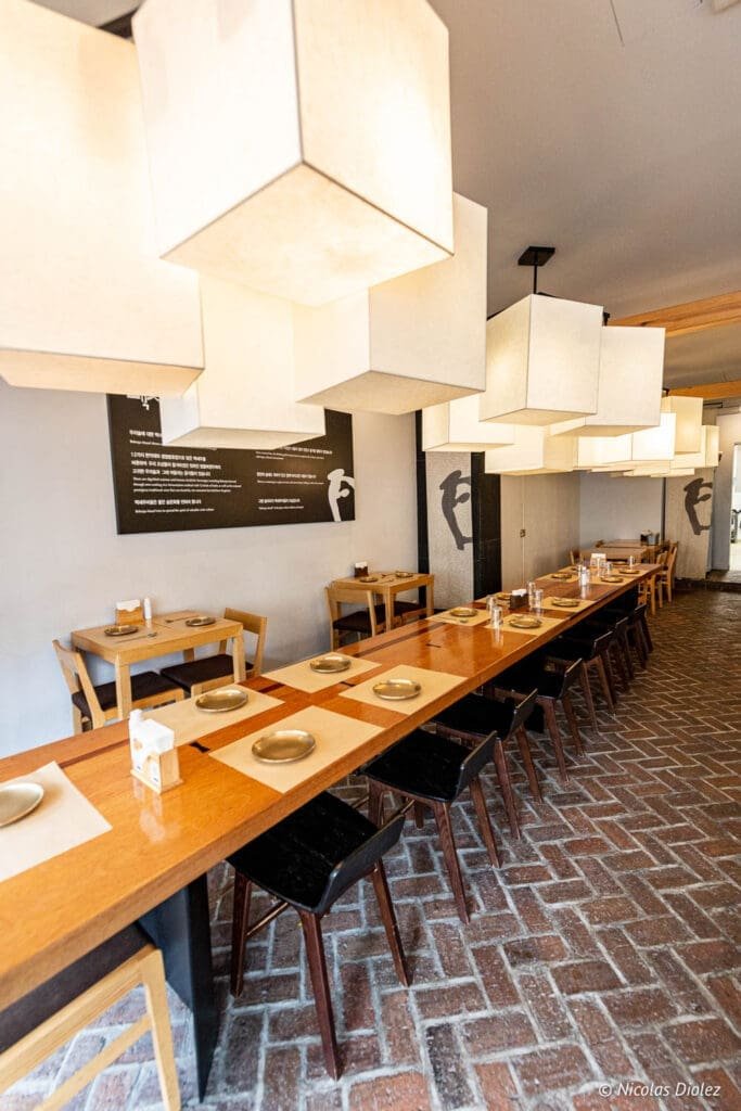 Intérieur du restaurant coréen Bekseju Village avec longue table en bois et luminaires cubiques.