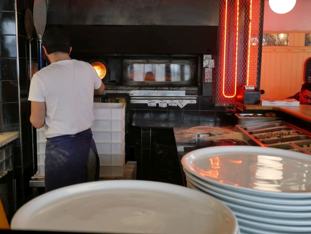 Pizzaiolo préparant une pizza devant un four dans une cuisine avec des assiettes empilées.