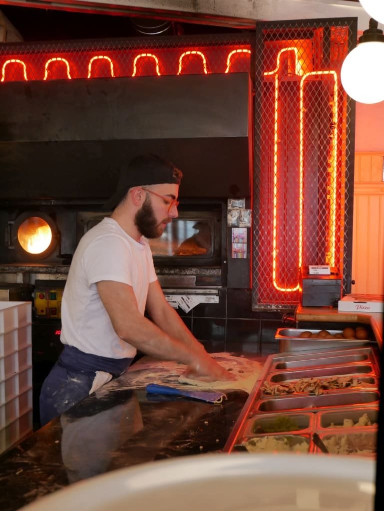 Pizzaiolo préparant la pâte à pizza devant un four et des néons rouges.