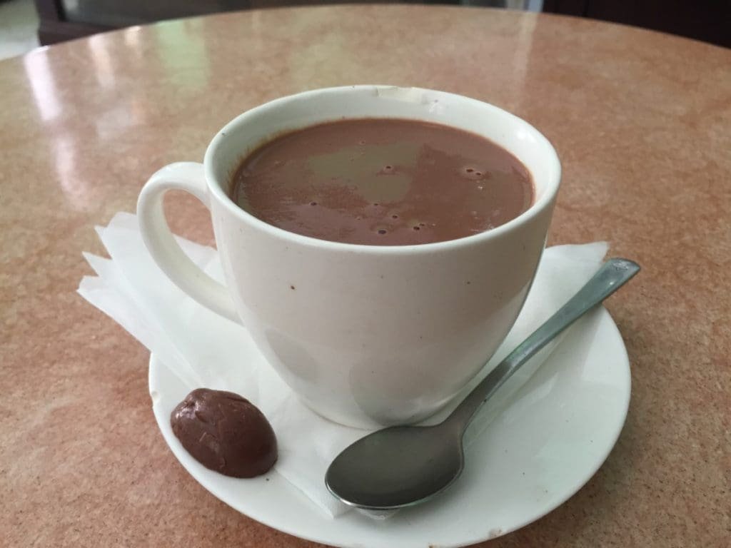 Musée du Chocolat. DR Valérie Collet 2018 Chocolat chaud dans tasse blanche, servi avec une cuillère et une petite friandise.