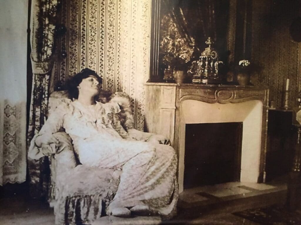 Lucy Hesse -DR Valérie Collet 2018 Portrait d'une femme assise en fauteuil devant une cheminée décorée.