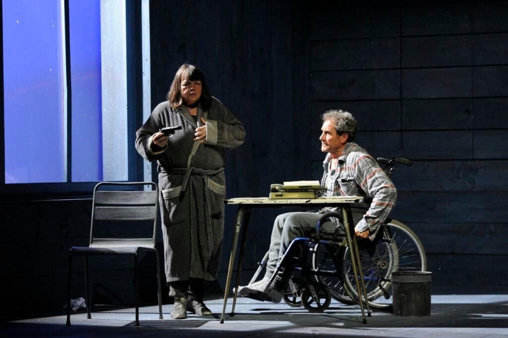 Misery theater scene: woman with a gun and man in a wheelchair typing.