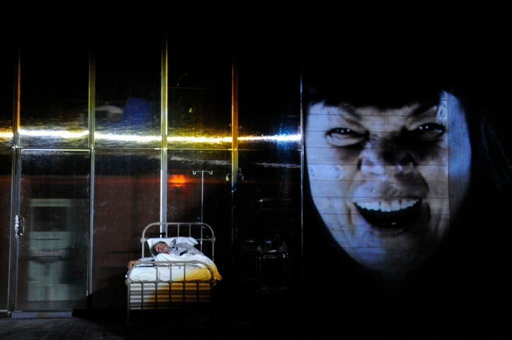 "Misery" theater scene: man in bed facing a threatening projected face.