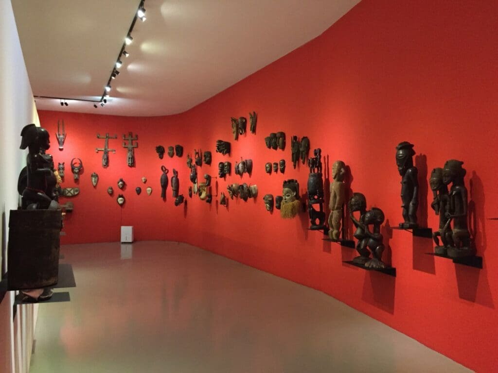 Aliança Underground Museum DR Valérie Collet 2019 Exposition masques et statues africains sur mur rouge, L'Aliança Underground Museum.