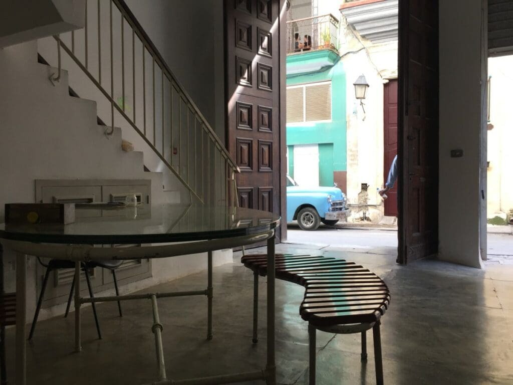 Factoria Habana DR Valérie Collet 2019 Intérieur cubain avec table ronde en verre et voiture ancienne bleue visible dehors.