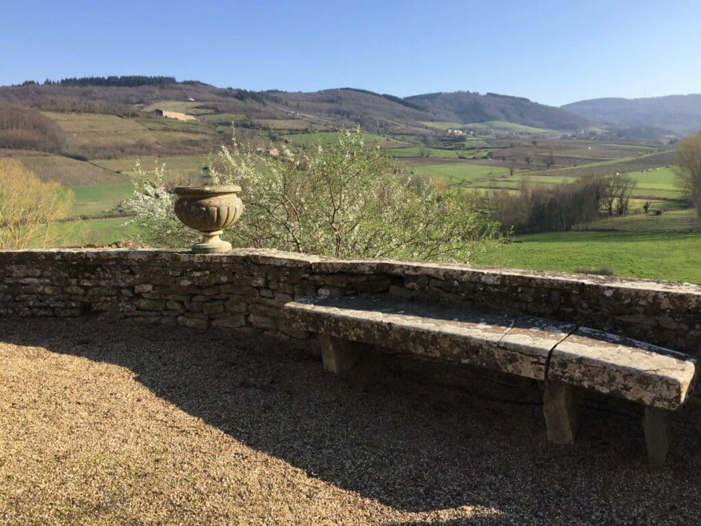 Château de Pierreclos - DR Valérie Collet 2019 Banc de pierre et urne sur terrasse avec vue sur les collines verdoyantes.