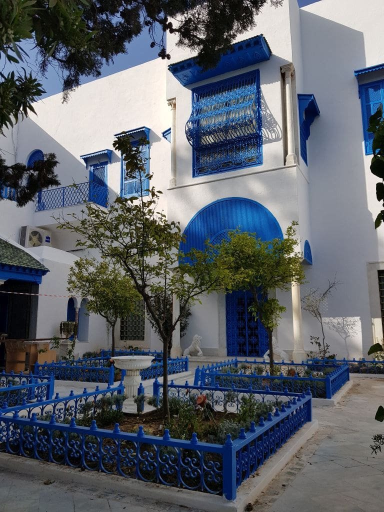 Palais Ennejma Ezzahra Sidi Bou Saïd - DR Melle Bon Plan 2019 Courtyard with white building, blue trim, and iron fence in Sidi Bou Saïd.