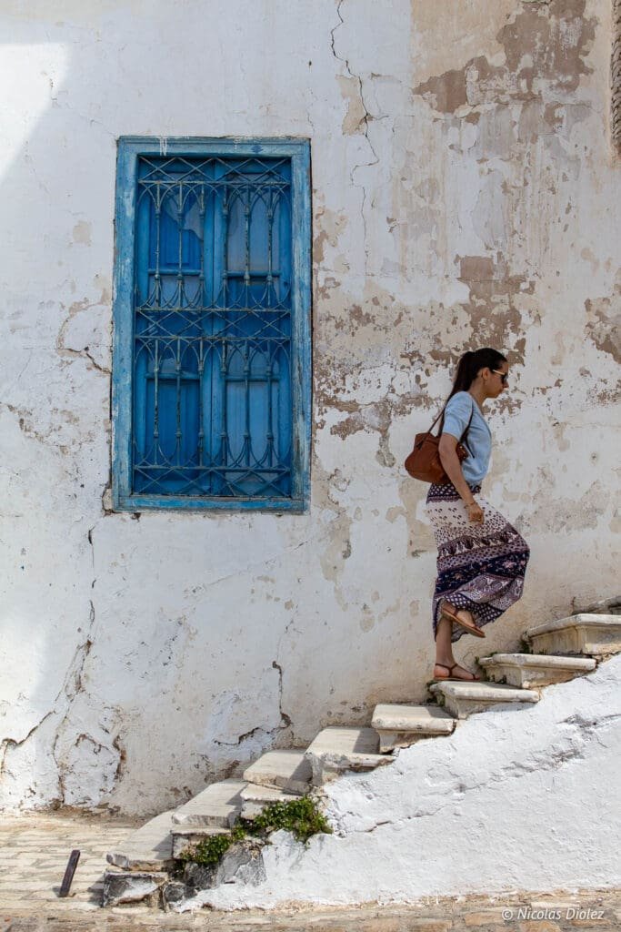 Femme montant un escalier blanc près d'une fenêtre bleue à Sidi Bou Saïd.
