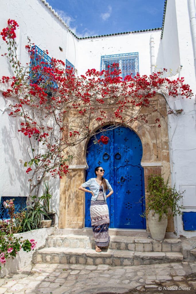 Femme devant porte bleue emblématique de Sidi Bou Saïd et bougainvilliers rouges.