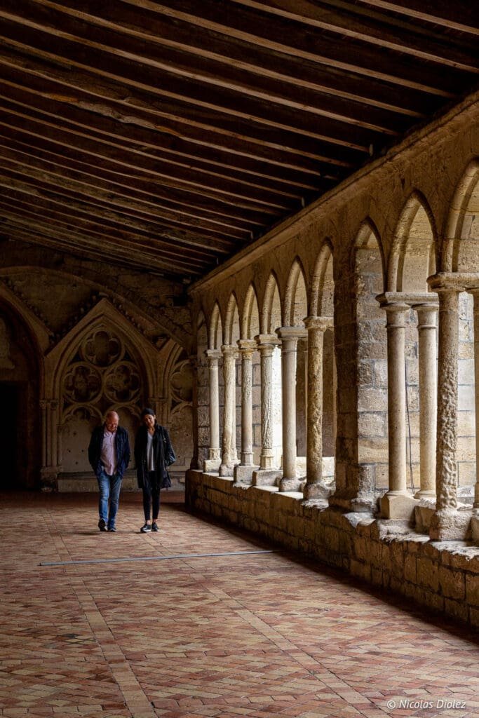 Cloister of Saint-Émilion with two visitors under a stone arch and wooden ceiling.