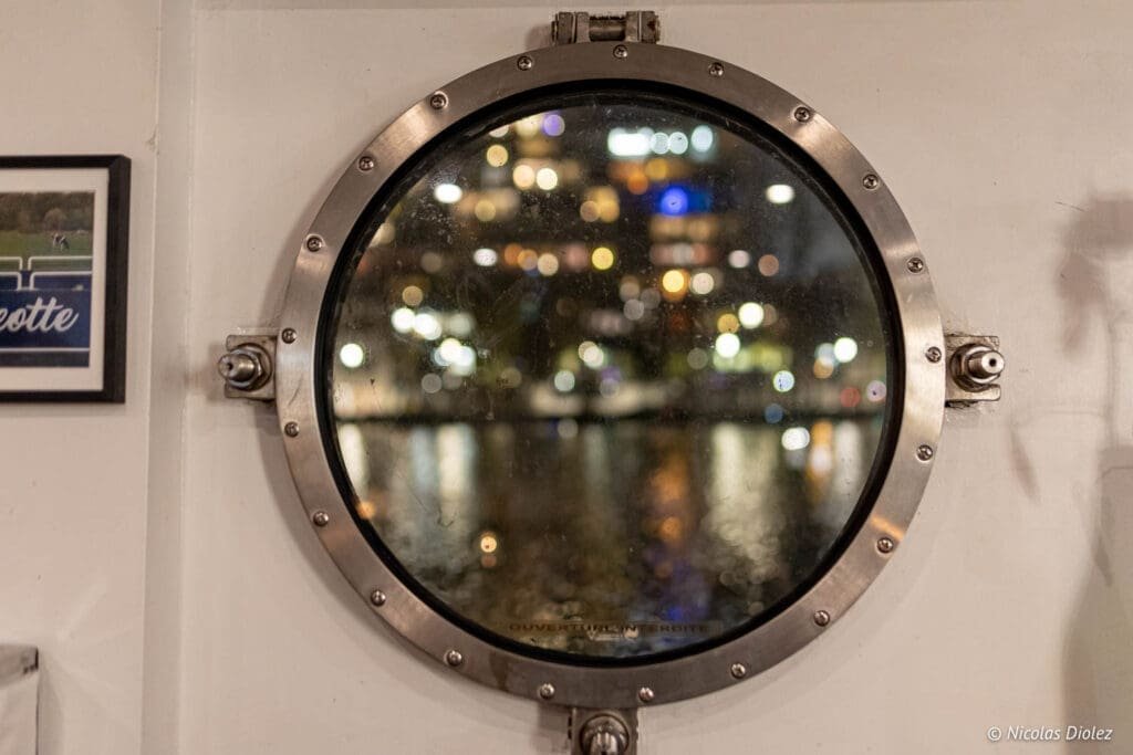 Hublot péniche La Bougeotte, vue nocturne urbaine avec reflets.