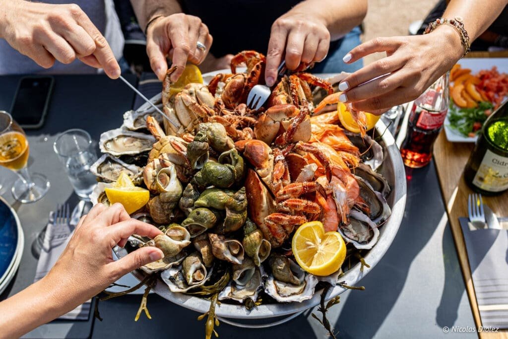 Plateau de fruits de mer O' P'tit Crabe de Goury avec mains dégustant huîtres et crevettes.