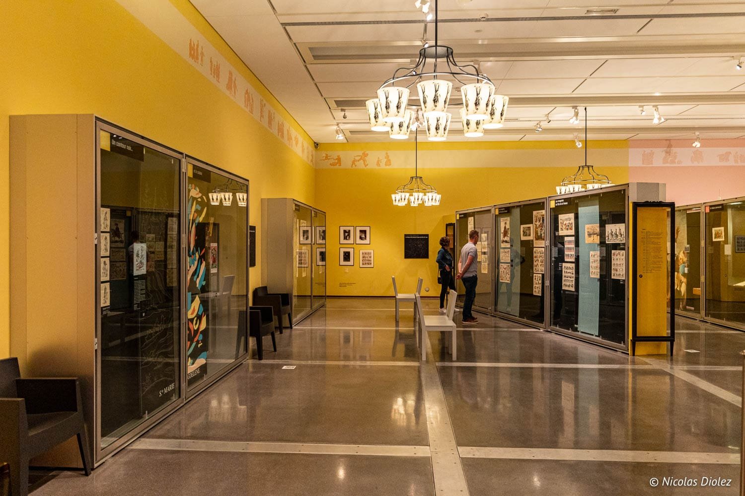 Intérieur jaune du Musée de l'Image d'Épinal avec visiteurs devant les vitrines d'estampes.