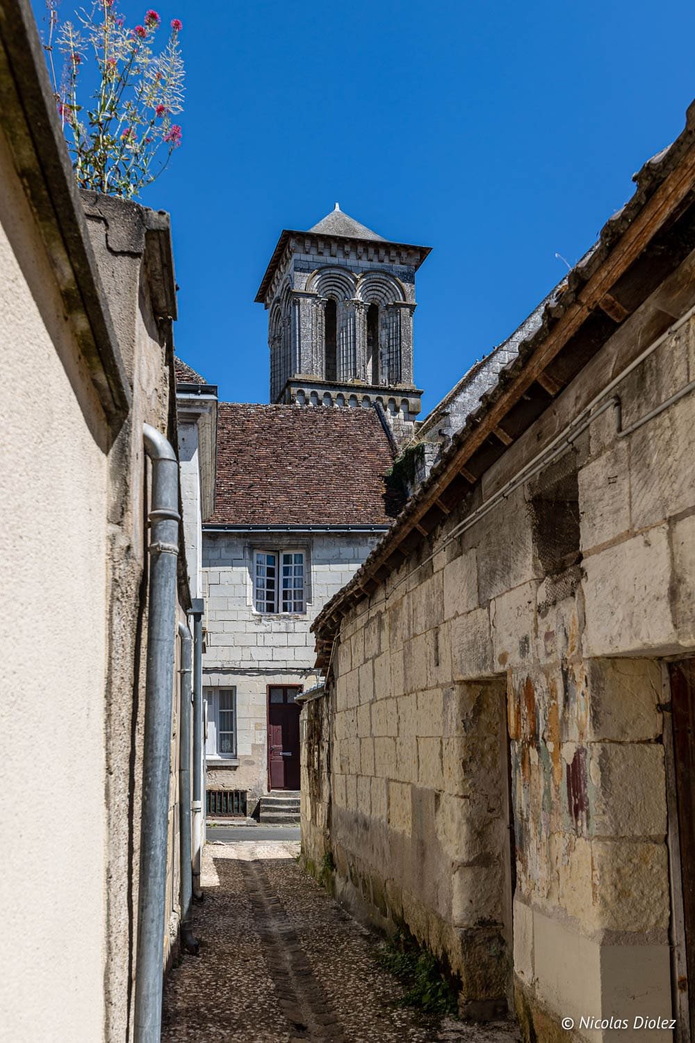 Ruelle pavée à Beaulieu-lès-Loches menant à un clocher roman sous le ciel bleu.
