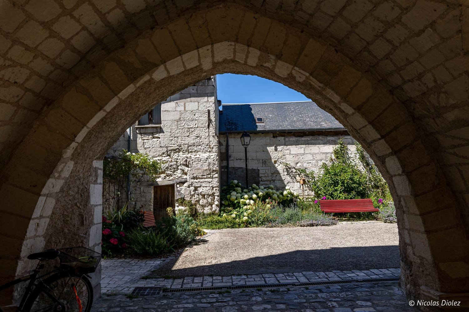 Passage voûté en pierre menant à une cour fleurie et banc rouge à Beaulieu-lès-Loches.