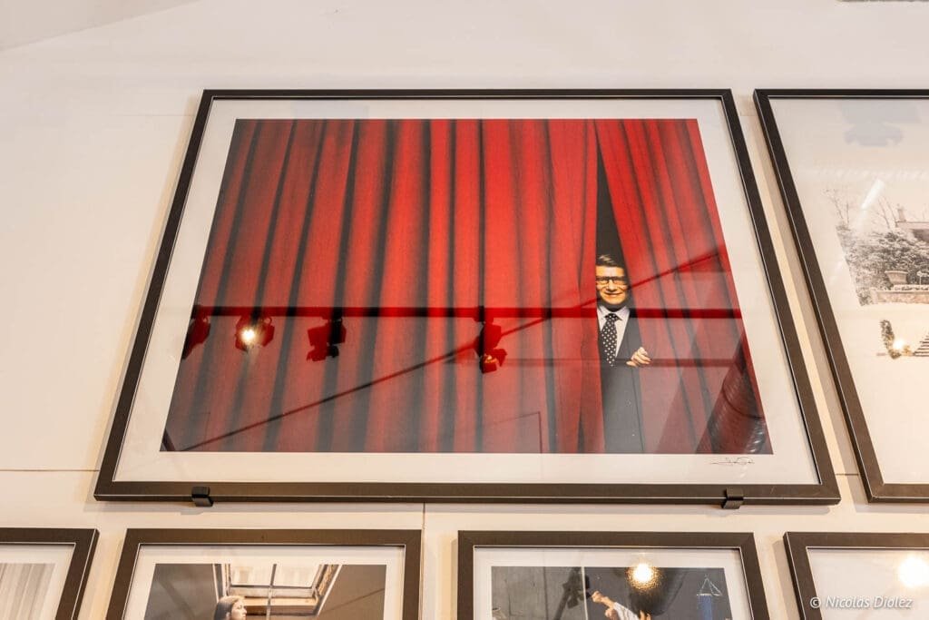 Exposition Jean-Marie Périer Villeneuve d'Aveyron, homme souriant derrière rideau rouge.