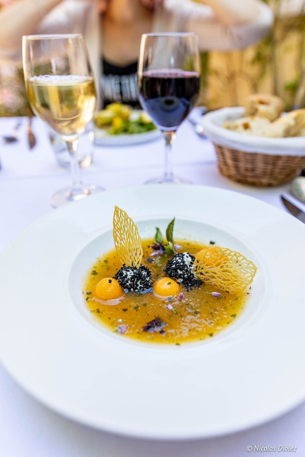 Assiette gourmande avec bouillon jaune, billes noires sésame, et vin blanc/rouge à table.