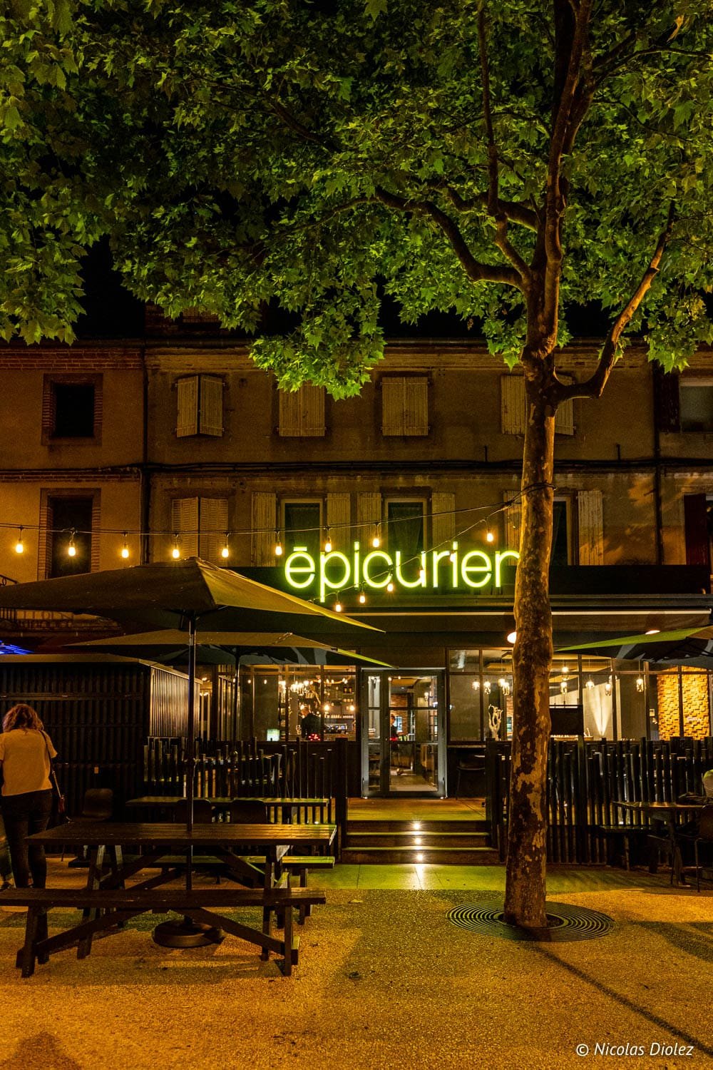 Façade du restaurant Épicurien la nuit avec terrasse et arbre central.