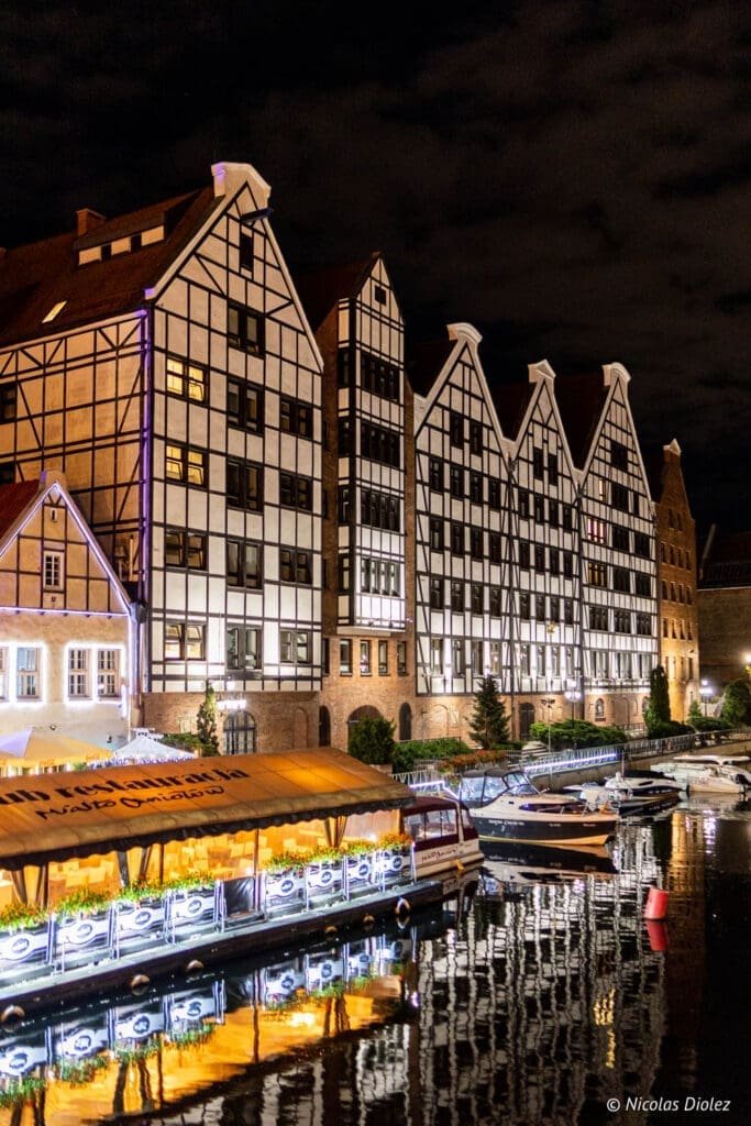 Bâtiments à colombages de Gdańsk la nuit avec reflets sur l'eau et bateaux.