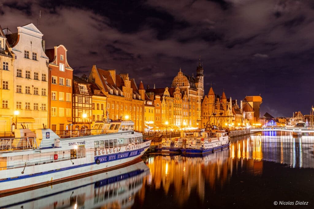 Quais de Gdańsk illuminés la nuit avec bateaux et reflets sur l'eau.