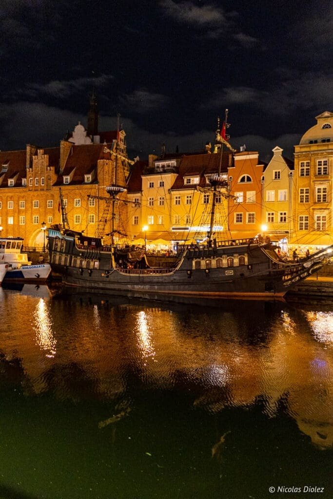 Gdańsk de nuit : galion pirate amarré sur la rivière Motława devant des maisons illuminées.