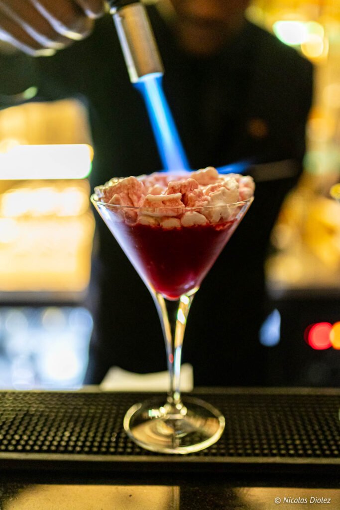 Barman che fiammeggia con un cannello la guarnizione di un cocktail rosso al bar dell'hotel Paris j'Adore.