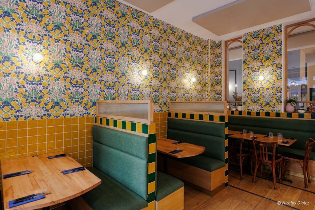 Intérieur restaurant avec banquettes vertes, tables bois, et papier peint fleuri jaune et bleu.