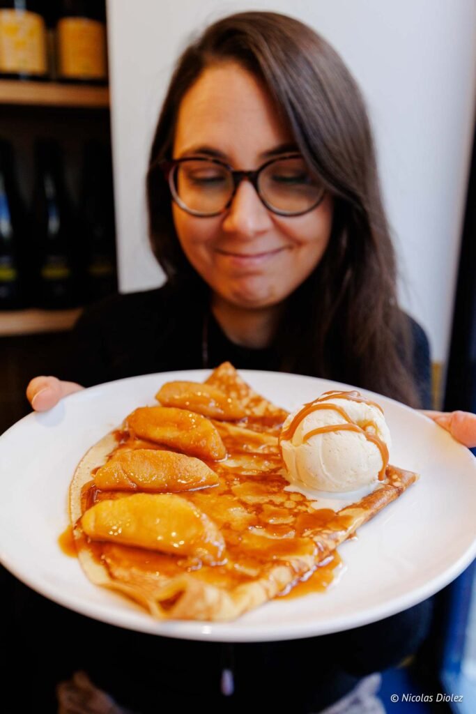 Crêpe aux pommes caramélisées et glace vanille tenue par femme souriante.
