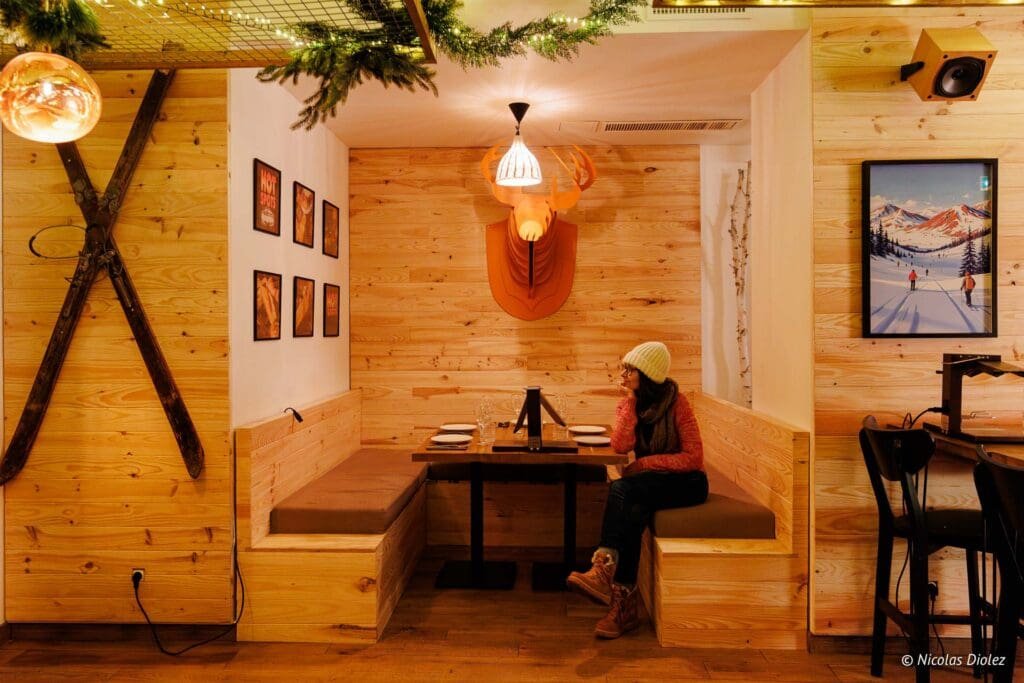 Intérieur chalet bois, femme seule assise à table, tête de cerf murale, skis.
