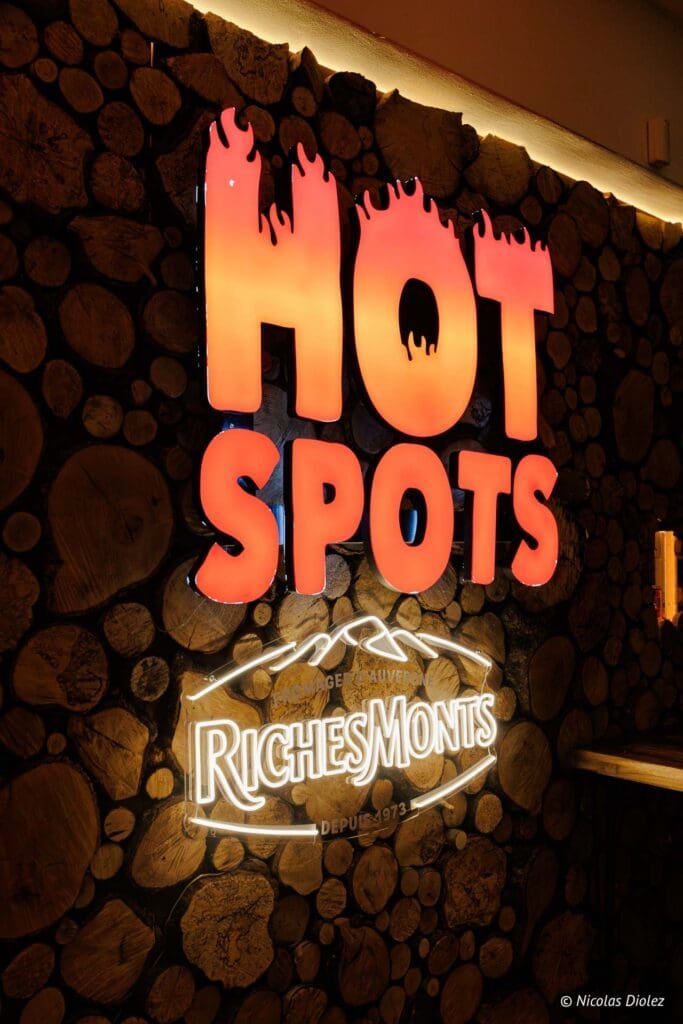 Enseigne lumineuse HOT SPOTS Richesmonts sur mur en rondins de bois.