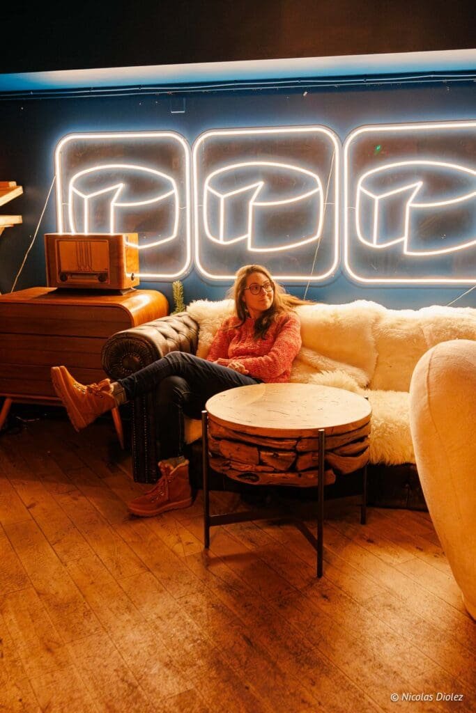 Femme assise sur canapé devant enseigne néon style fromage, intérieur cosy.