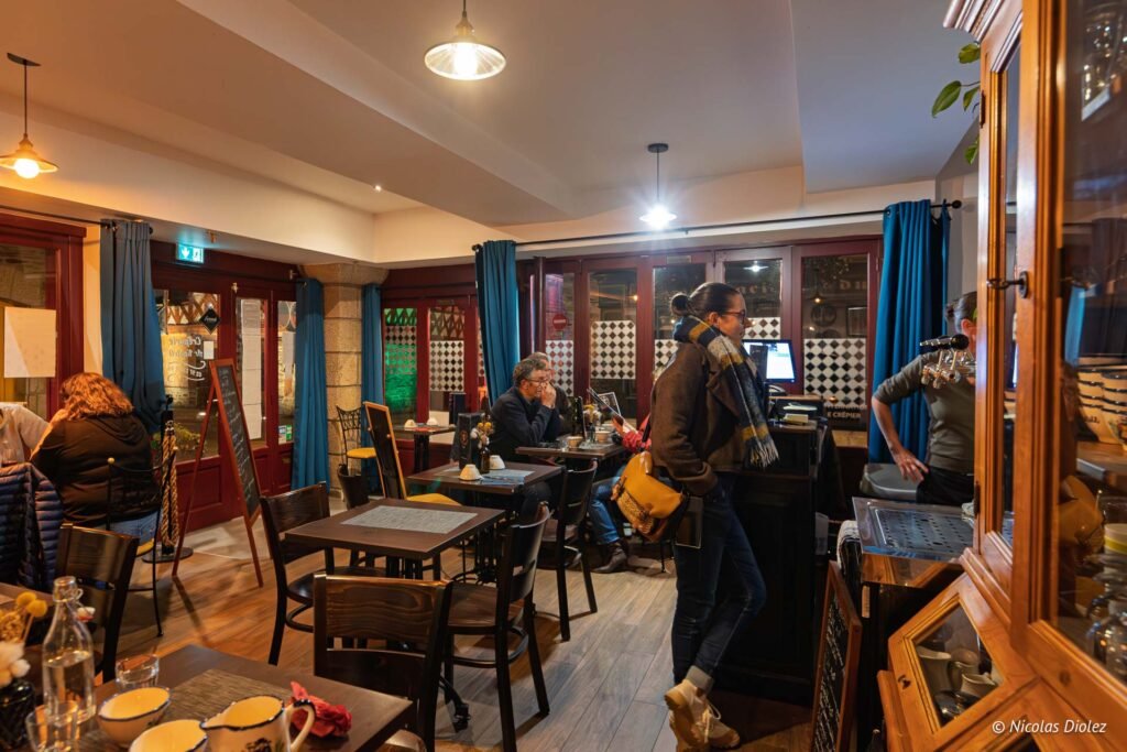 Intérieur restaurant crêperie, clients attablés près des fenêtres, femme debout près du comptoir.