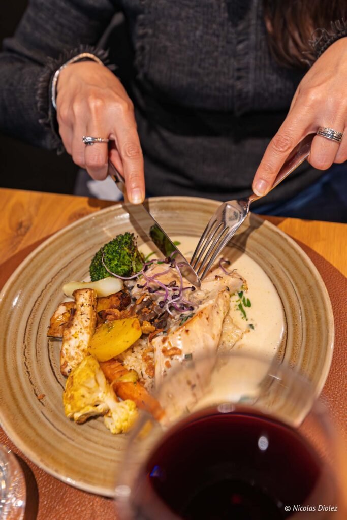Personne coupant un plat de viande avec légumes et sauce crémeuse, verre de vin.