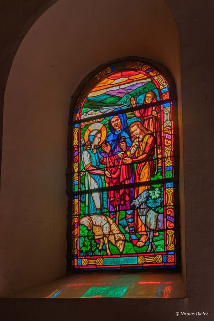 Vitrail de l'église du Graal montrant la Vierge, un homme en bleu et un autre en rouge avec des moutons.