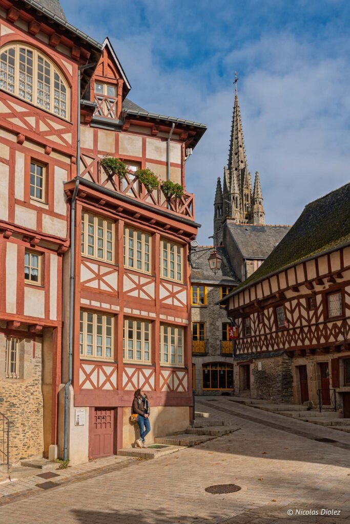 Rue pavée Josselin avec maisons à pans de bois et flèche d'église en arrière-plan.