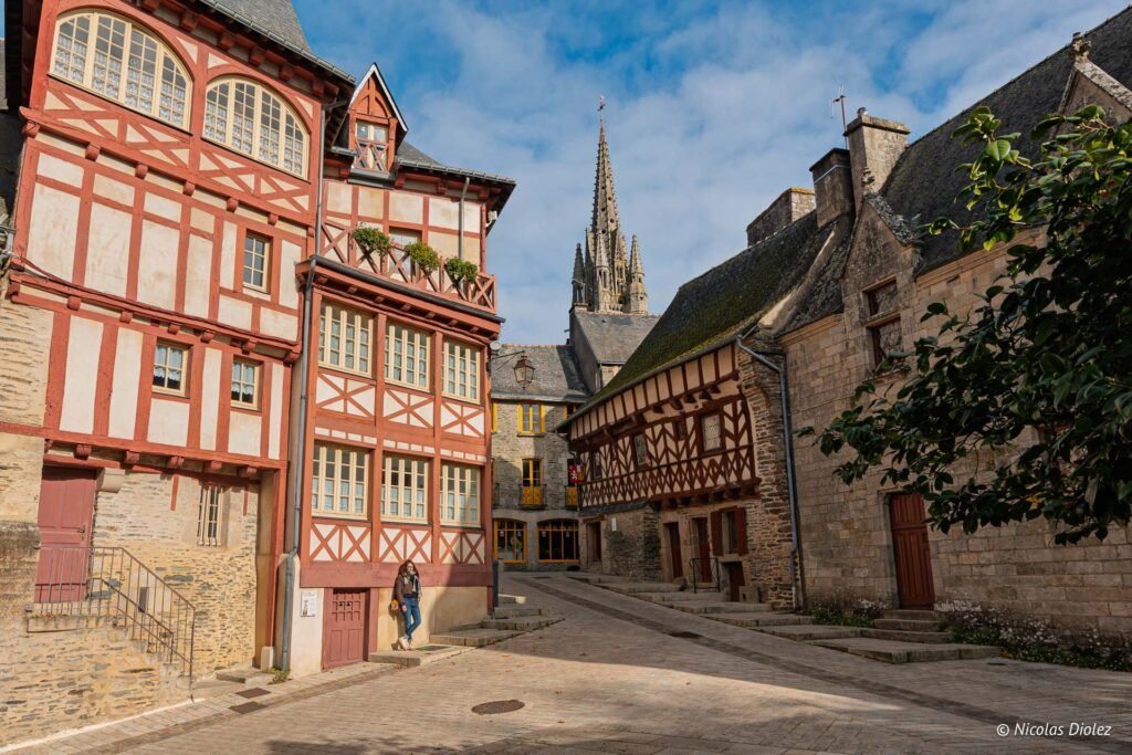 Rue pavée à Josselin avec maisons à pans de bois et flèche d'église.