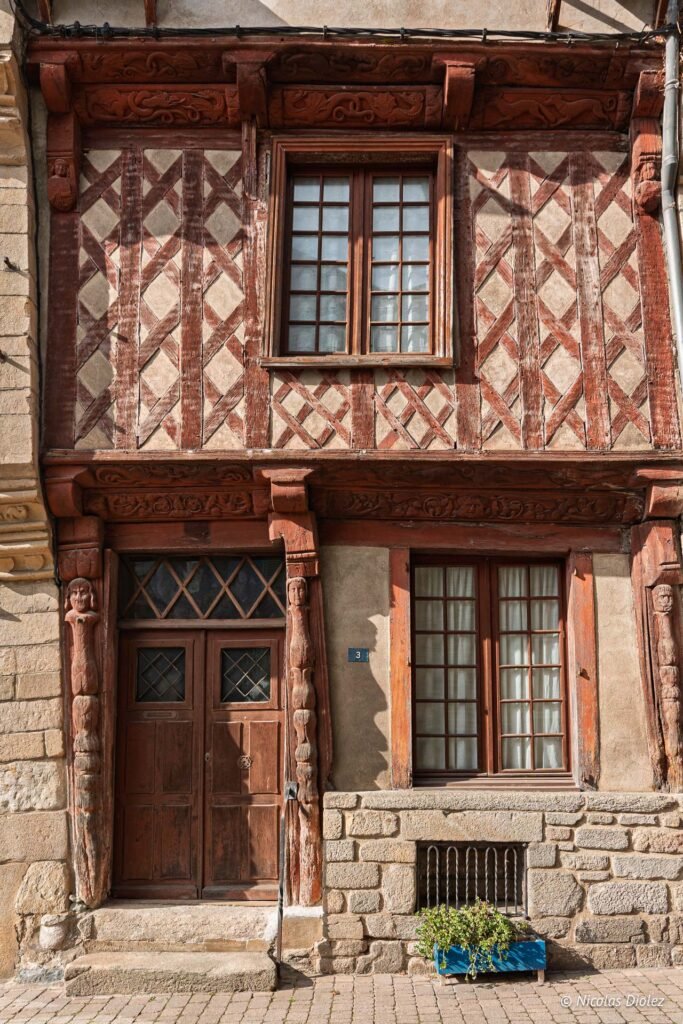 Façade à colombages rouges, porte en bois sculptée, et fenêtres à croisillons.