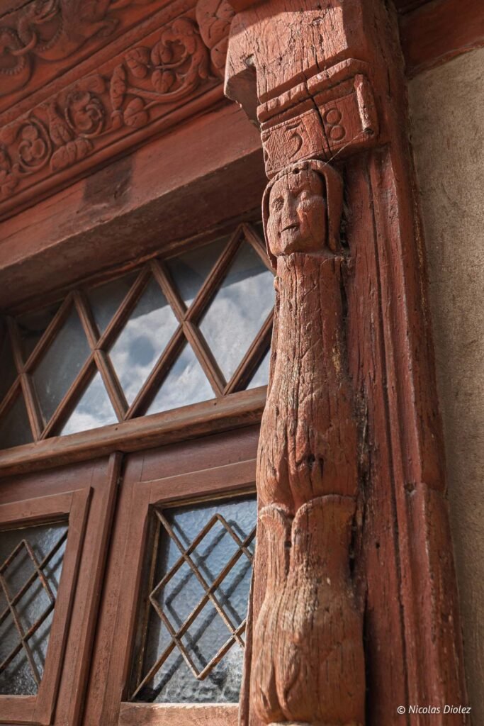 Poteau en bois sculpté avec visage et linteau sculpté, fenêtre à croisillons.