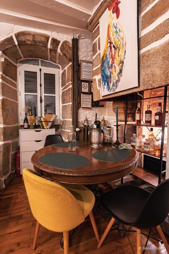 Coin repas rustique avec table ronde, tableau coq coloré et mur en pierre.