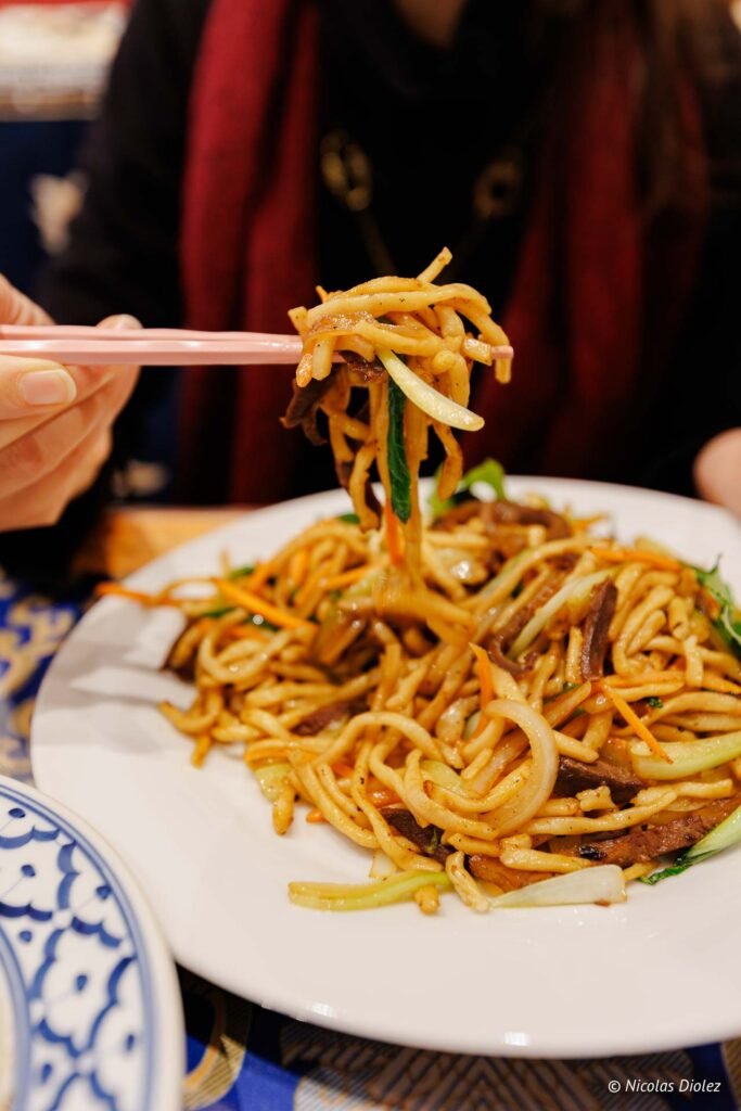 Mains de baguettes soulevant des nouilles sautées aux légumes et viande. Nicolas Diolez.