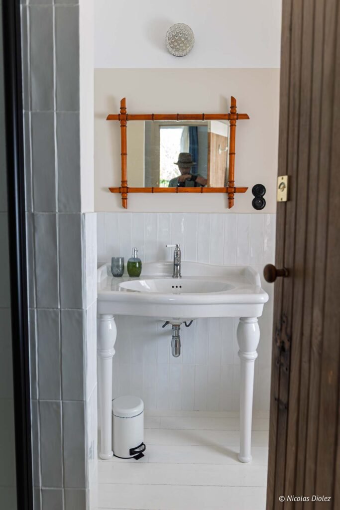 Salle de bain avec vasque blanche sur pieds, miroir cadre bambou et porte en bois.