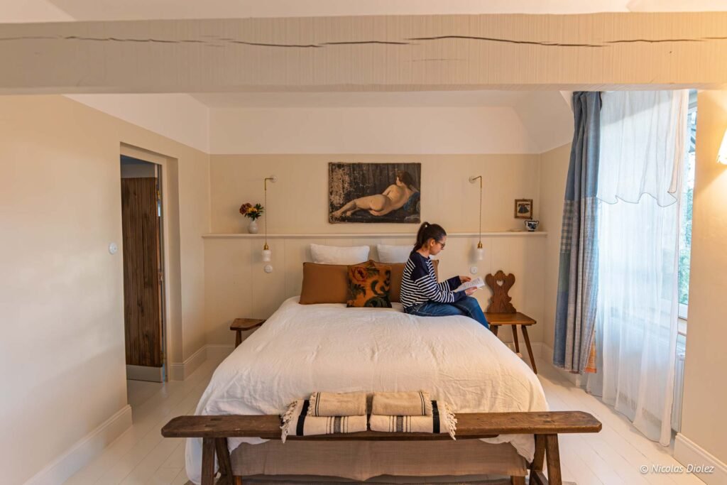 Femme lisant assise sur un grand lit blanc dans une chambre rustique, tableau nu.