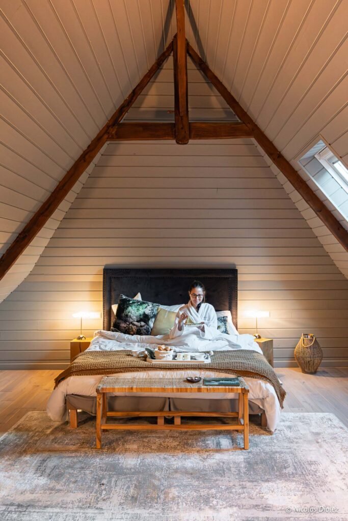 Femme en peignoir mange au lit sous toits mansardés Manoir du Vau d'Arz.