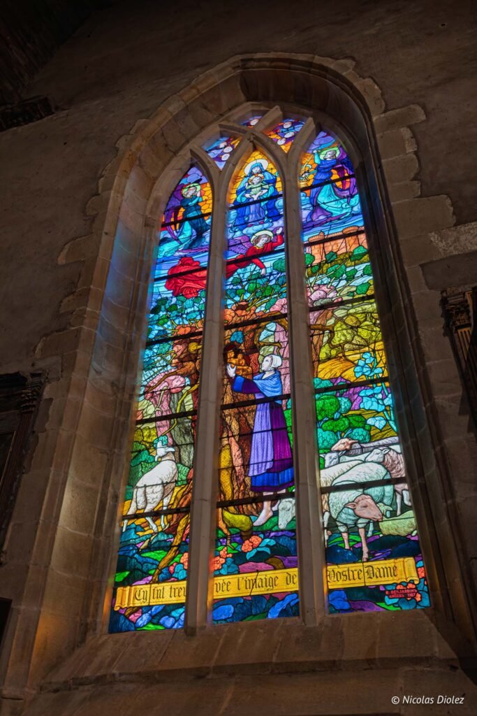 Vitrail coloré représentant une scène religieuse avec une femme, des moutons, et le texte "Nostre Dame".
