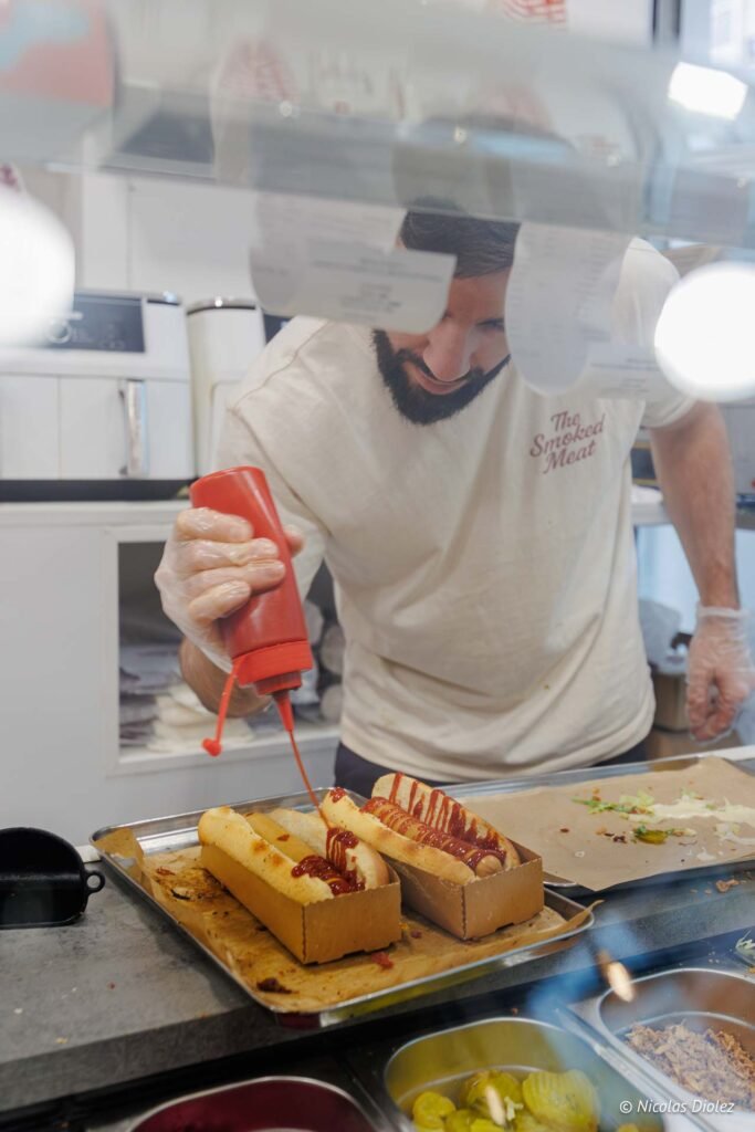 Employé de The Smoked Meat ajoutant du ketchup sur des hot-dogs.