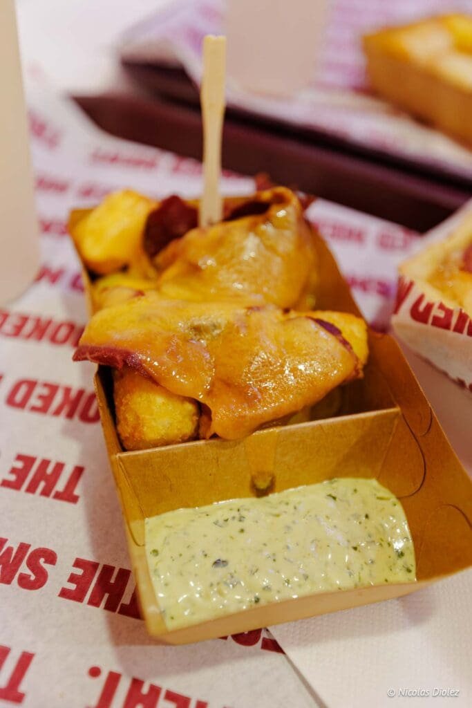 Frites nappées de fromage fondu et sauce verte dans un contenant en carton.